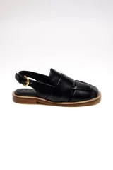 Sandalia negra de cuero con diseño tejido en el empeine, tira trasera con hebilla dorada ajustable y suela plana.