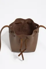 Bolso tipo bucket pequeño de piel vacuna, color marrón, con estructura blanda. Cuenta con asa regulable de piel y cierre de cordón ajustable. Presenta el logo metálico de BIMBA Y LOLA en dorado.