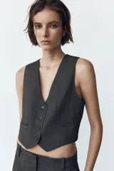 Chaleco corto gris oscuro con escote en pico, sin mangas, bolsillos de ojal y cierre frontal con botones.
