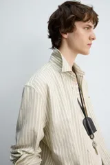 Camisa de hombre relaxed fit confeccionada en tejido de algodón a rayas verticales color beige y marrón. Tiene cuello solapa, manga larga con puños abotonados, bolsillo de parche en el pecho y cierre frontal con botones.