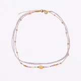 Collar de doble capa con cadena dorada y cordón trenzado lila/blanco. La capa inferior presenta cuentas de piedras semipreciosas en tonos pastel y una piedra central ovalada de color amarillo/naranja, con detalles de cuentas doradas en la capa superior.