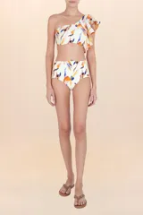 Top de bikini asimétrico, color blanco hueso, con estampado abstracto en tonos naranja, azul y rosado. Presenta un volado amplio en el hombro derecho y tirantes finos ajustables.