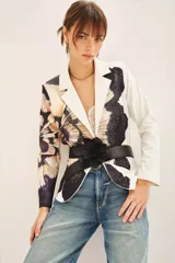 Blazer con estampado abstracto en tonos beige, negro y blanco. Tiene solapas blancas y está adornado con tachas termofijadas en el frente. Se cierra con un cinturón negro con una flor grande.