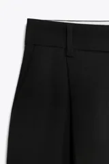 Pantalón negro de tiro alto con trabillas, pinzas delanteras, bolsillos laterales y falsos bolsillos de vivo en la espalda. Cierre frontal con cremallera y botón.
