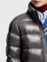 Campera puffer corta de plumas Moncler modelo Maya en color gris con acabado brillante. Presenta un diseño acolchado con capucha, cierre frontal de cremallera, bolsillos laterales y un pequeño bolsillo con solapa y parche del logo en la manga izquierda.