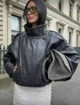 Chaqueta negra de efecto cuero, estilo bomber o motociclista, con cuello alto abotonado y cierre frontal asimétrico. Presenta detalles de costuras verticales y hombreras pequeñas. El bajo es elástico.