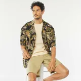 Camisa de manga corta con estampado de hojas tropicales en tonos verdes, marrones y rosados sobre fondo negro.