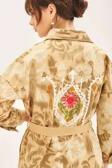 Campera de corte recto confeccionada en gabardina estampada con bordados de hilo. Presenta cuello camisero, cierre frontal con botones, dos bolsillos de parche con solapa en el pecho y lazo ajustable en la cintura con detalle bordado.