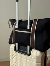 Bolso tipo tote de neopreno color negro y marrón, con cierre superior, bolsillo frontal marrón con logo rojo, bolsillo interno y externo con cierre, doble asa de mano marrón con detalles blancos y opción de correa larga.