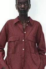 Camisa color borgoña a rayas verticales finas, de corte recto, con cuello solapa y manga larga. Tiene dos bolsillos de parche con solapa en el pecho y cierre frontal con botones.