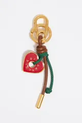 Llavero con dije de corazón rojo esmaltado con logo grabado, cordón de cuero vacuno marrón con nudo y cordón trenzado verde. Incluye un mosquetón metálico en tono dorado para colgar.