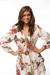 Vestido corto blanco con estampado floral en tonos rojos, verdes y amarillos. Tiene escote en V, mangas largas abullonadas y falda con volados. Se ajusta a la cintura con un cinturón de soga con hebilla circular.