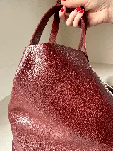 Cartera tipo tote bag color bordó, confeccionada en cuero con textura granulada. Tiene doble asa corta de mano y asa larga extraíble con diseño de tachas para usar como bandolera.