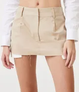 Minifalda color beige de sarga con cintura baja, trabillas, cierre con corchete y cremallera, y forro de bolsillo aparente.