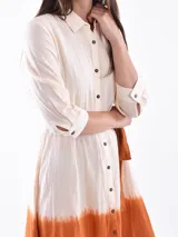 Vestido midi color crudo con efecto degradado color naranja en la parte inferior. Tiene corte camisero, cuello clásico, mangas 3/4 y cinturón de tela color naranja.