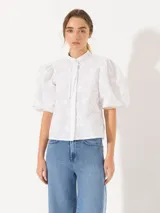 Camisa blanca de poplin con cuello mao y lazo para atar. Tiene mangas cortas abullonadas con flecos en los puños y cierre frontal oculto.