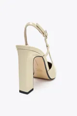 Zapato de tacón destalonado color beige acharolado, con punta fina, tacón ancho en bloque y cierre mediante tira trasera con hebilla ajustable.