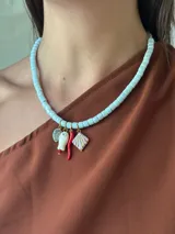 Collar corto tipo gargantilla compuesto por cuentas cilíndricas de cerámica color celeste pálido. Incluye dijes colgantes: una hoja verde, un amuleto blanco con detalle rojo, un chile rojo y una caracola blanca con detalles rosados.