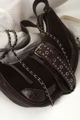 Cartera bandolera de gamuza color chocolate con detalles en cuero liso y cuero con textura croco. Presenta una correa de cuero para llevar al hombro y una cadena metálica desmontable. La solapa frontal está decorada con tachas metálicas y tiene cierre imantado.