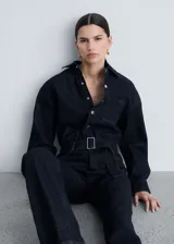 Mono largo de denim azul oscuro, con cuello camisero, mangas largas con puños abotonados, trabillas, cinturón con hebilla metálica, cierre frontal con botones a presión y cuatro bolsillos.