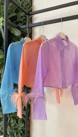 Camisa corta de mangas largas, con cuello y botones, confeccionada en tela traslúcida color lila y terminaciones de plumas en los puños.