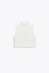 Chaleco blanco acolchado con cuello alto y cierre frontal con cremallera. Tiene bolsillos delanteros con cierre de cremallera oculta y bajo ajustable con cordones elásticos laterales.