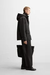 Parka negra de corte regular, confeccionada en tejido técnico acolchado. Tiene cuello alto con capucha ajustable, mangas largas con puños ajustables, bolsillos con solapa en la cadera y un bolsillo interior. El cierre frontal es con cremallera oculta por solapa con botones a presión.