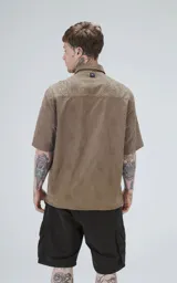 Camisa de manga corta color verde militar con cuello camisero y botones frontales oscuros. La tela presenta un patrón texturizado o estampado sutil en tono similar.