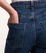 Pantalón mom confeccionado en jeans azul con trabillas en la cintura, cierre tradicional por botón y cremallera, bolsillos delanteros y traseros con detalle deshilachado.