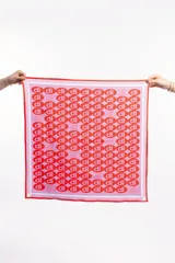 Pañuelo cuadrado de seda o material similar, con patrón repetitivo de las letras 'dl' en círculos. El fondo es rosa claro/lavanda y los círculos son de color rojo/coral, con un borde rojo sólido.