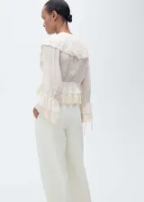 Blusa color crema con cuello en V, volados y detalles de encaje. Tiene mangas largas con puños elásticos y lazos.