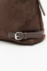 Cartera de gamuza color marrón, de textura suave y acabado aterciopelado. Cuenta con una silueta amplia y flexible, cierre superior metálico, correa regulable y detalles laterales con hebillas.