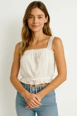 Top corto color blanco crudo, con escote cuadrado y tirantes anchos de encaje. Presenta detalles de broderie vertical y un cordón ajustable en el ruedo inferior.