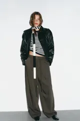 Chaqueta negra de efecto piel con cuello solapa, manga larga y trabillas en los hombros. Presenta bolsillos delanteros y cierre frontal con botones, ajustada a la cintura con un cinturón del mismo tejido.