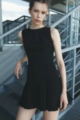 Vestido corto negro sin mangas, con cuello redondo y falda con tablas. Cierre trasero con cremallera oculta.