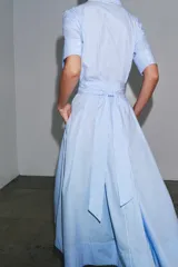 Vestido camisero midi de popelín a rayas verticales celestes y blancas, con cuello solapa, manga corta, bolsillos de plastrón con solapa en el pecho, cinturón de lazo ajustable y cierre frontal con botones.
