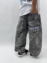 Pantalón de jean gris con efecto lavado, corte holgado y tiro alto. Presenta bolsillos delanteros y traseros, y estampados con letras en la parte trasera.