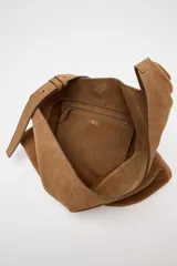 Bolso tipo saca asimétrica de serraje color marrón. Tiene asa bandolera ajustable y cierre con imán. Incluye cartera con bolsillo extraíble.
