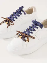 Zapatillas urbanas de cuero vacuno color blanco, con interior al tono y talonera en descarne color beige. Presentan doble cordón, uno azul y otro marrón, y dijes colgantes.