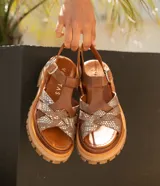 Sandalias bajas de cuero vacuno color beige con tiras cruzadas plateadas y hebilla lateral. Suela doble de goma deportiva con altura de 3cm adelante y 5,5cm atrás.