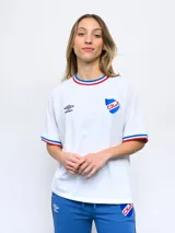 Remera blanca de corte clásico con cuello redondo y mangas cortas con puños tricolor (azul, blanco y rojo). Presenta el logo de Umbro bordado en el pecho izquierdo y el escudo del Club Nacional de Football bordado en el pecho derecho.