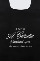 Bolso tipo shopper negro con asas cortas. Estampado frontal con el logo de Zara, la leyenda "A Coruña Established 1975" y la dirección "Rúa Juan Flórez 64-66" en color blanco.