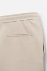 Bermuda color beige de corte recto, con cintura elástica ajustable con cordón, detalle de pliegues frontales, bolsillos laterales y bolsillos de vivo en la espalda.