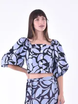 Blusa corta con estampado de hojas en tonos celeste y negro, escote cuadrado, mangas abullonadas 3/4 y cierre frontal con botones blancos.