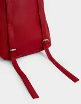 Mochila pequeña de cuero sintético color blanco con bolsillo frontal con solapa y cierre superior. Incluye un charm colgante de moño a rayas rojas y blancas con dije de corazón plateado.