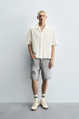 Bermuda de denim de algodón, color gris lavado, con corte relaxed fit y roturas visibles en la parte delantera de ambas piernas. Presenta cinco bolsillos y cierre frontal con cremallera y botón.