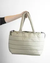 Bolso tipo tote color beige con diseño acolchado y asas tubulares. Presenta el logo de la marca en el centro.