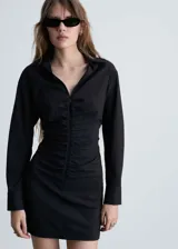 Vestido corto camisero negro con finas rayas verticales blancas, cuello camisero, mangas largas con puños abotonados, cierre frontal con cremallera y frunces verticales en el centro.