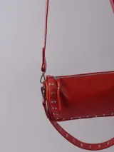 Bandolera color rojo de cuero con tachas plateadas. Tiene dos asas desmontables, una corta con tachas para llevarla en la mano o al hombro y una larga para llevarla al hombro o cruzada. Tiene bolsillo interior con cierre y forro.