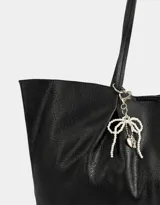 Cartera shopper negra de cuerina con manijas superiores y acceso mediante broche imantado. Incluye charm de perlas en forma de moño.